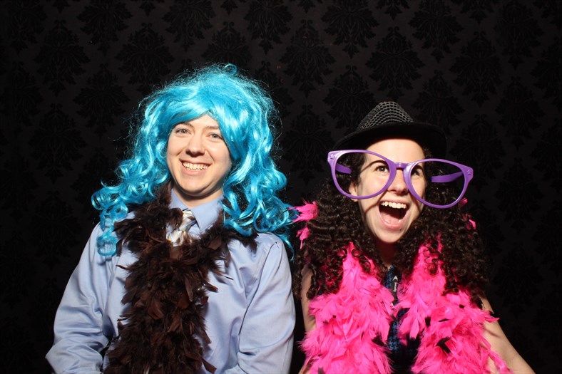 Ryan and Sharon at Photo Booth. Ryan is wearing a blue haired wig, and Sharon has on a bright pink boa with purple sunglasses.