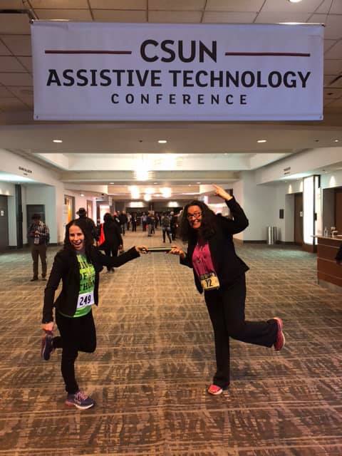 Sharon and Dana, both wearing race bibs, and passing a baton under the CSUN Assistive Technology sign. 